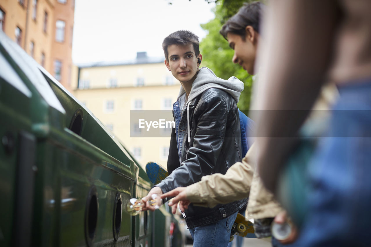 Teenage boy looking at friend while throwing waste in garbage bin at recycling station