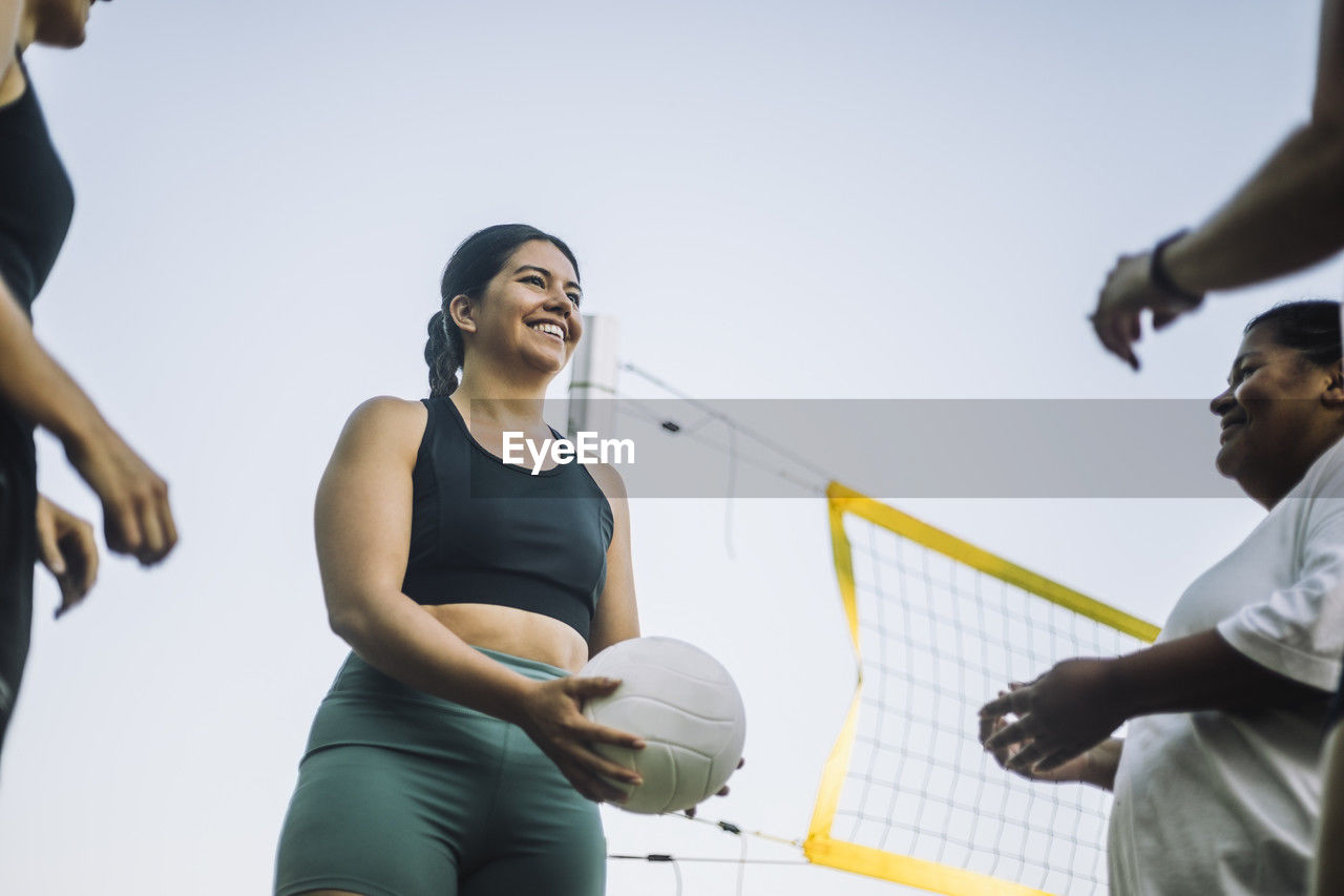 Low angle view of smiling woman playing volleyball with friends against sky