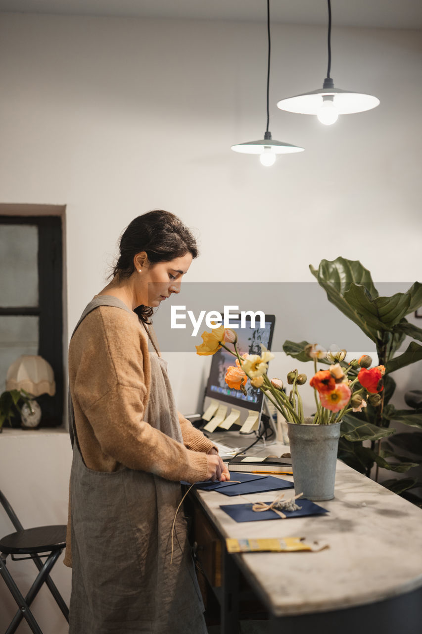 Diligent worker in brown sweater and gray apron concentrating and making decor elements for designing modern apartments while standing at table with computer and fresh peach color flowers in vase in contemporary flower shop