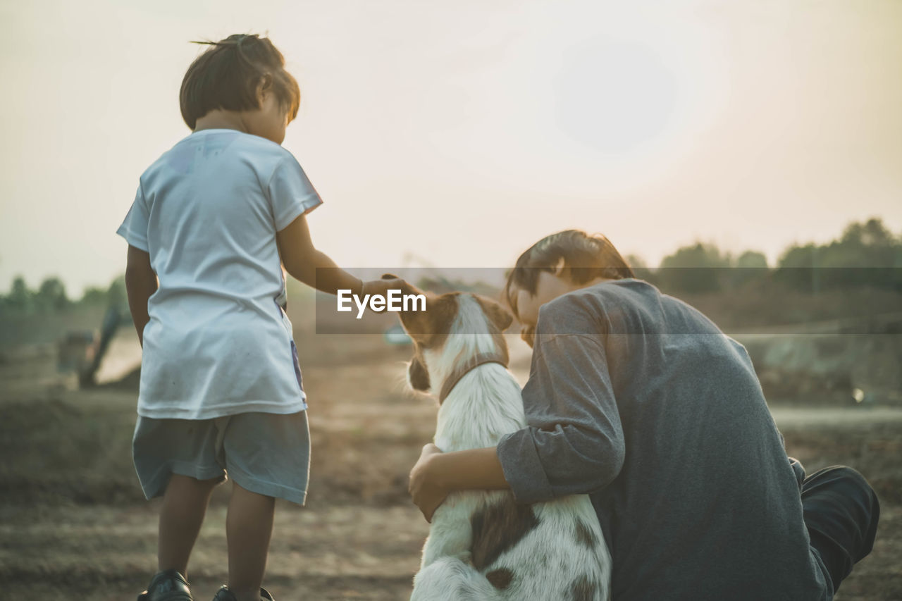 Rear view of mother and daughter with dog on field against clear sky during sunset