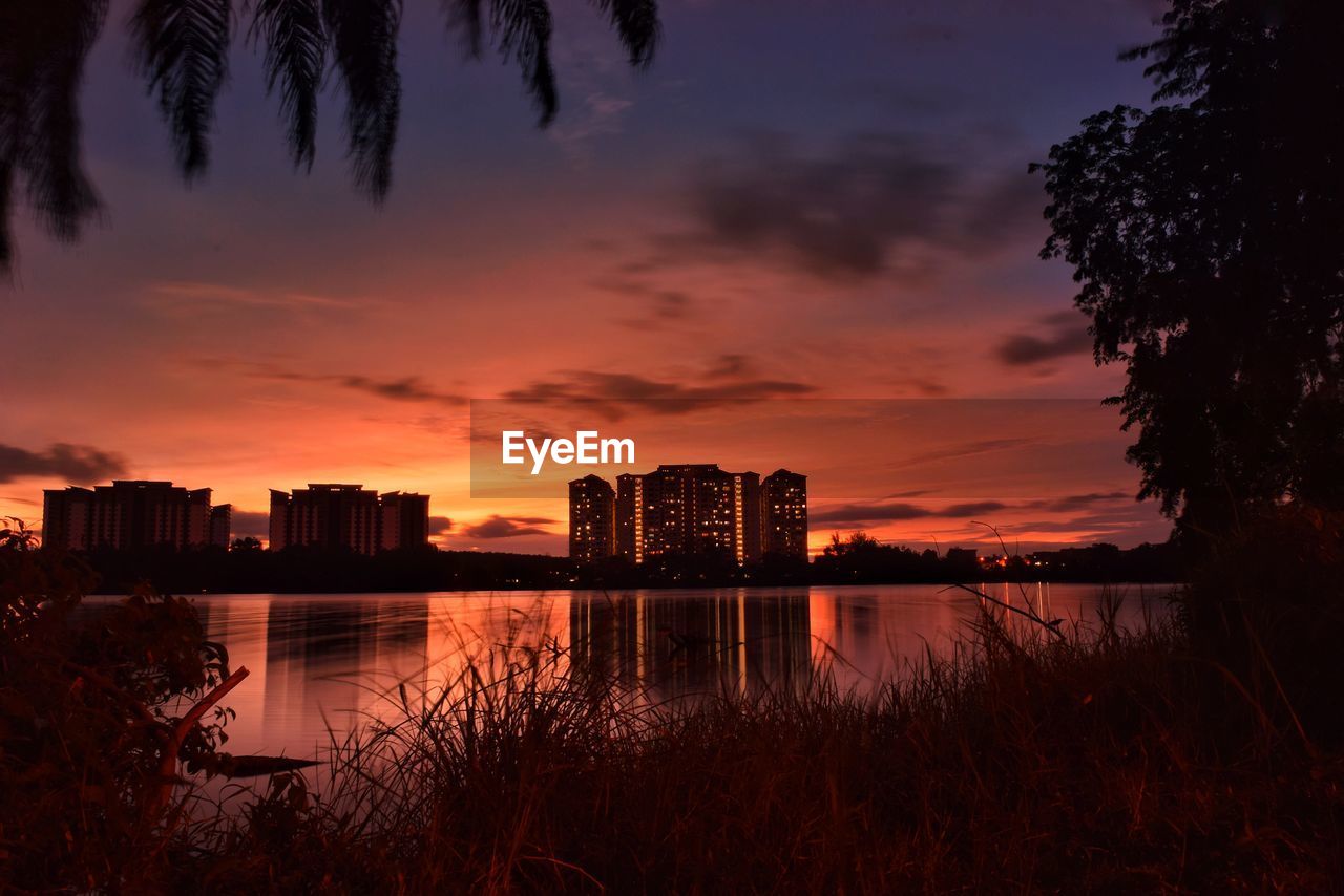 Silhouette buildings by lake against sky during sunset