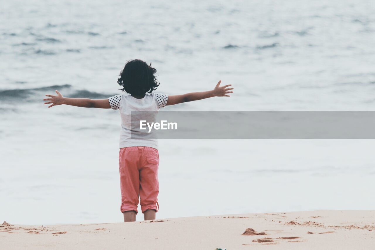 REAR VIEW OF GIRL STANDING ON BEACH AGAINST SKY