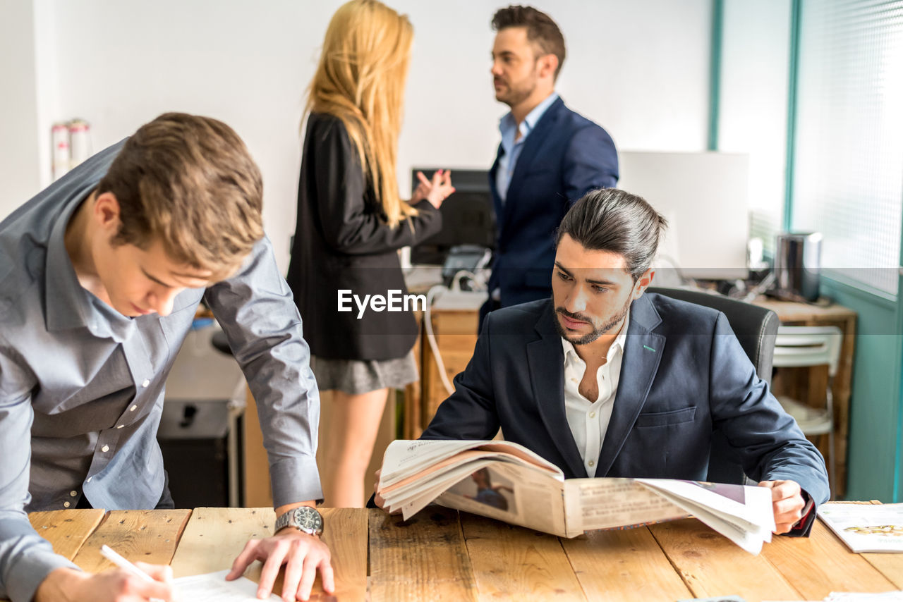 Focused male worker in formal wear taking notes in documents on table while working in light modern office with colleagues