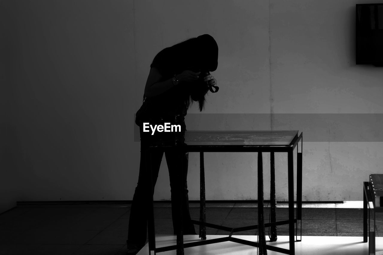 Woman photographing while standing by table against wall