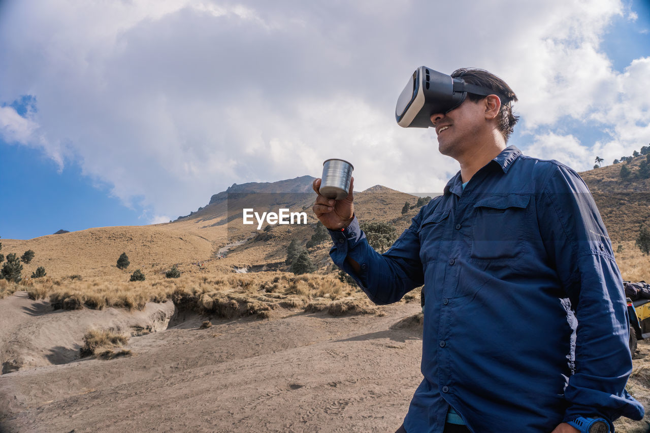 Man holding mug while wearing virtual reality headset against mountain