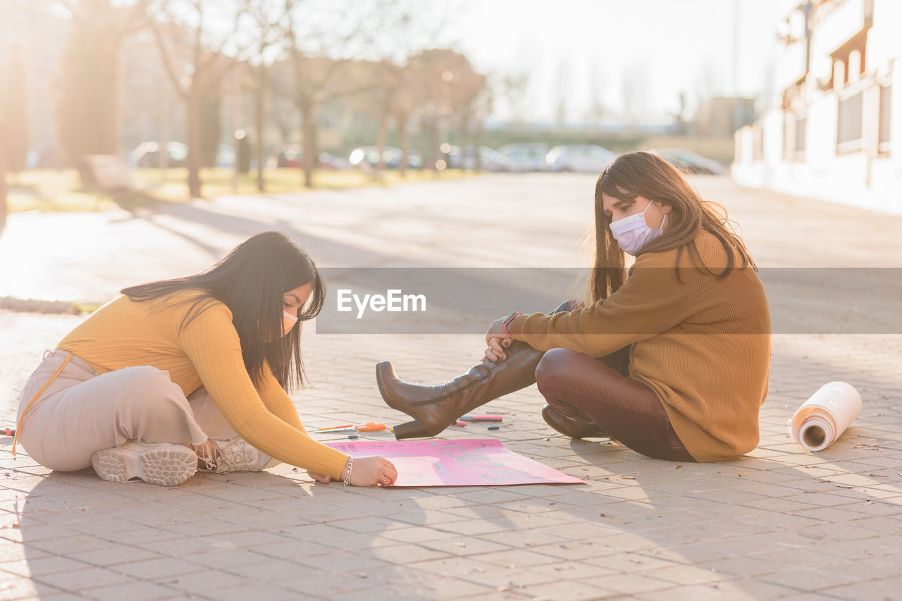 Females wearing mask writing on banner while sitting on street