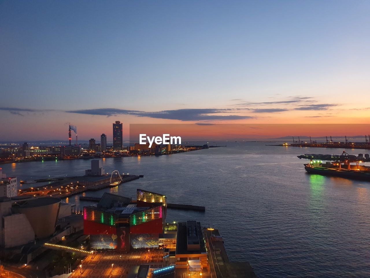 Illuminated city by sea against sky during sunset