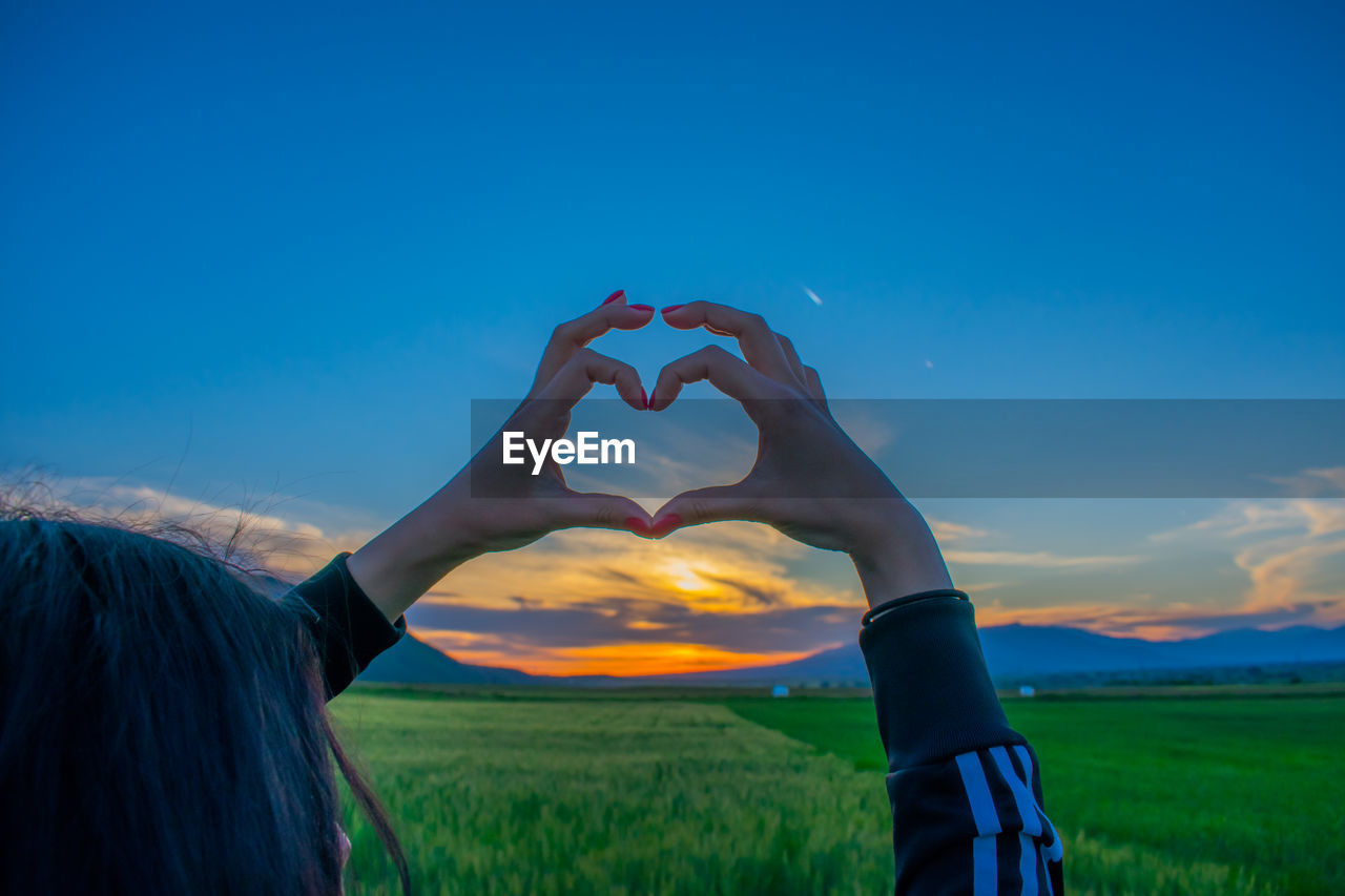 WOMAN HAND HOLDING HEART SHAPE ON FIELD AGAINST SKY DURING SUNSET