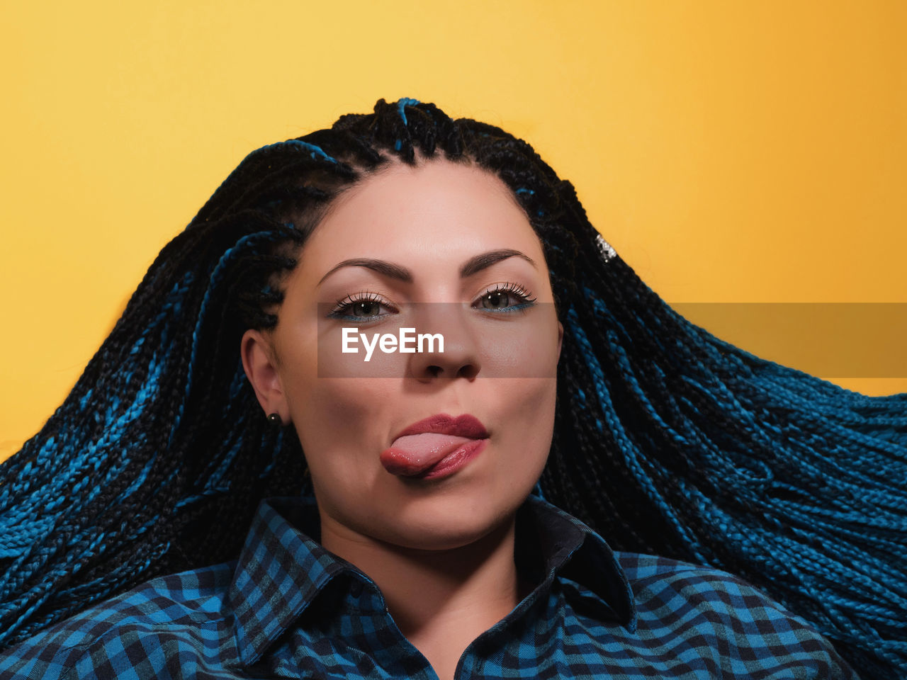 Portrait of young woman with braided hair sticking out tongue against yellow background