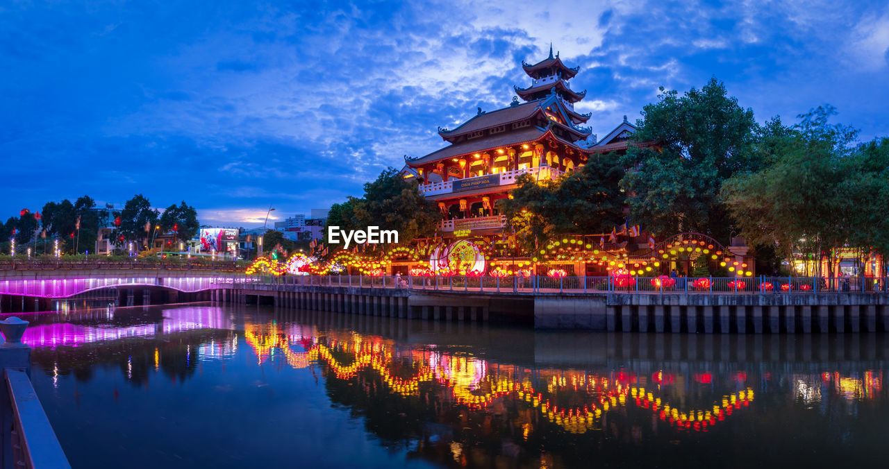 Illuminated building by lake against sky at dusk