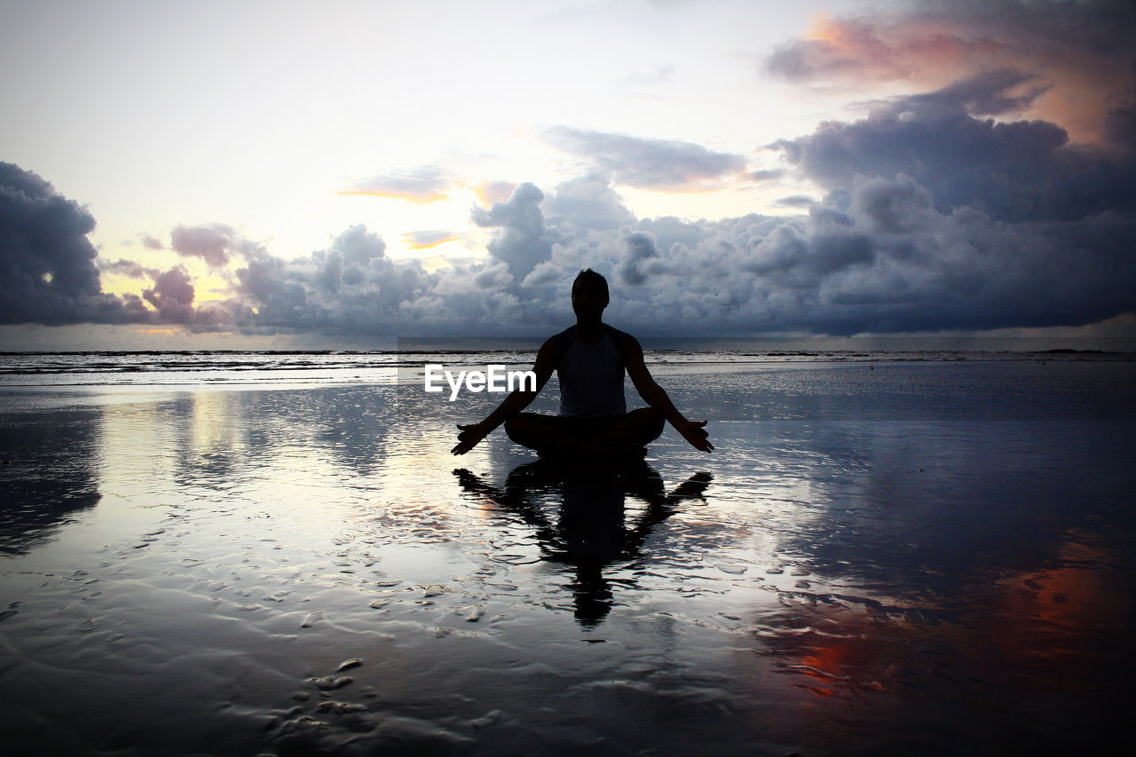 Silhouette man sitting in lotus position on shore at beach against sky