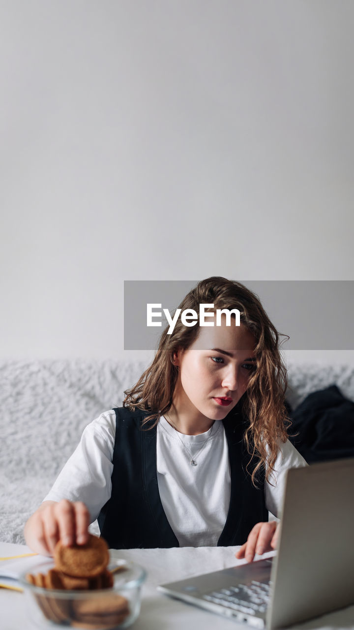 Young woman watching series or podcasts, looking at the screen with interest, holding cookies in