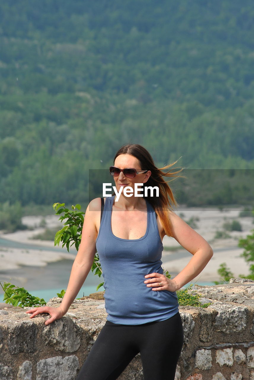 Smiling beautiful woman wearing sunglasses while standing against retaining wall