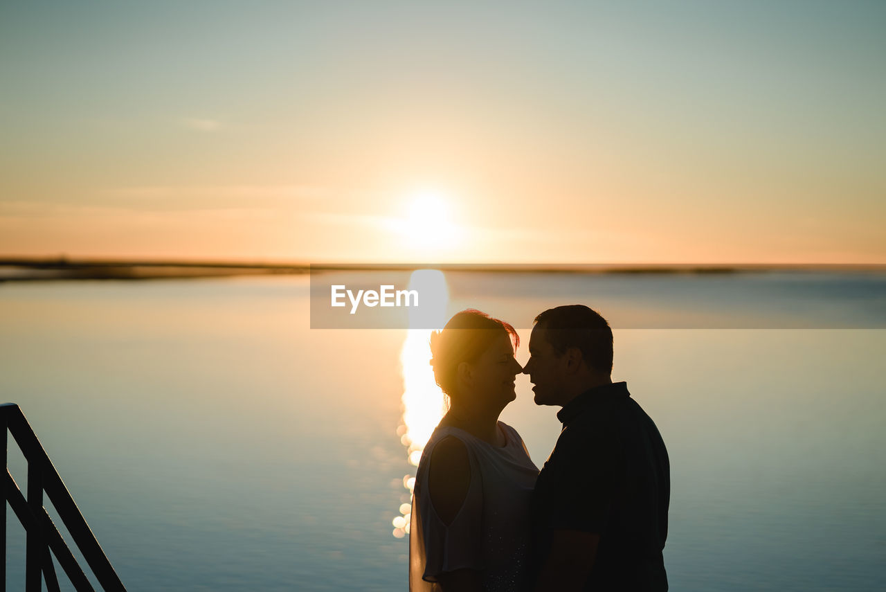 MAN AND WOMAN STANDING AT SEA DURING SUNSET