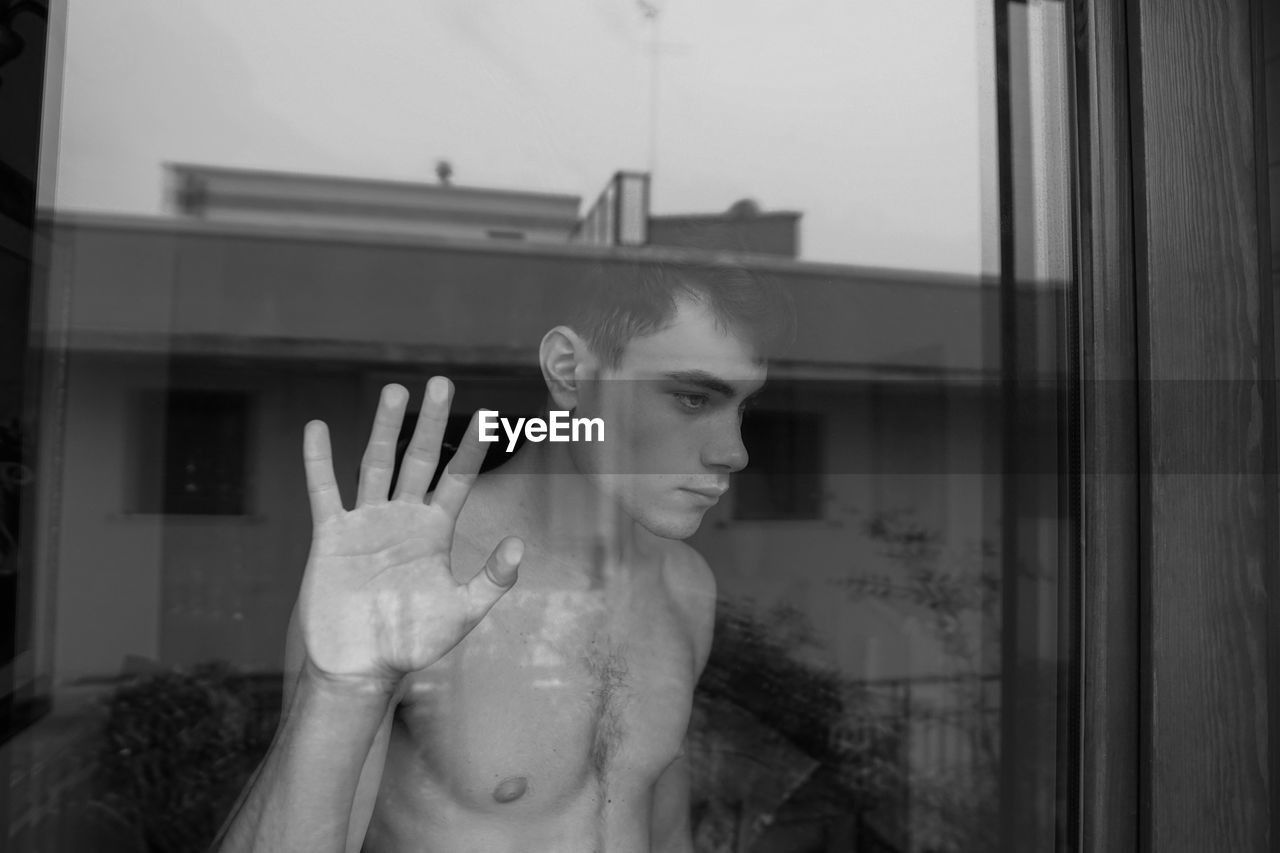 Thoughtful shirtless teenage boy looking through window