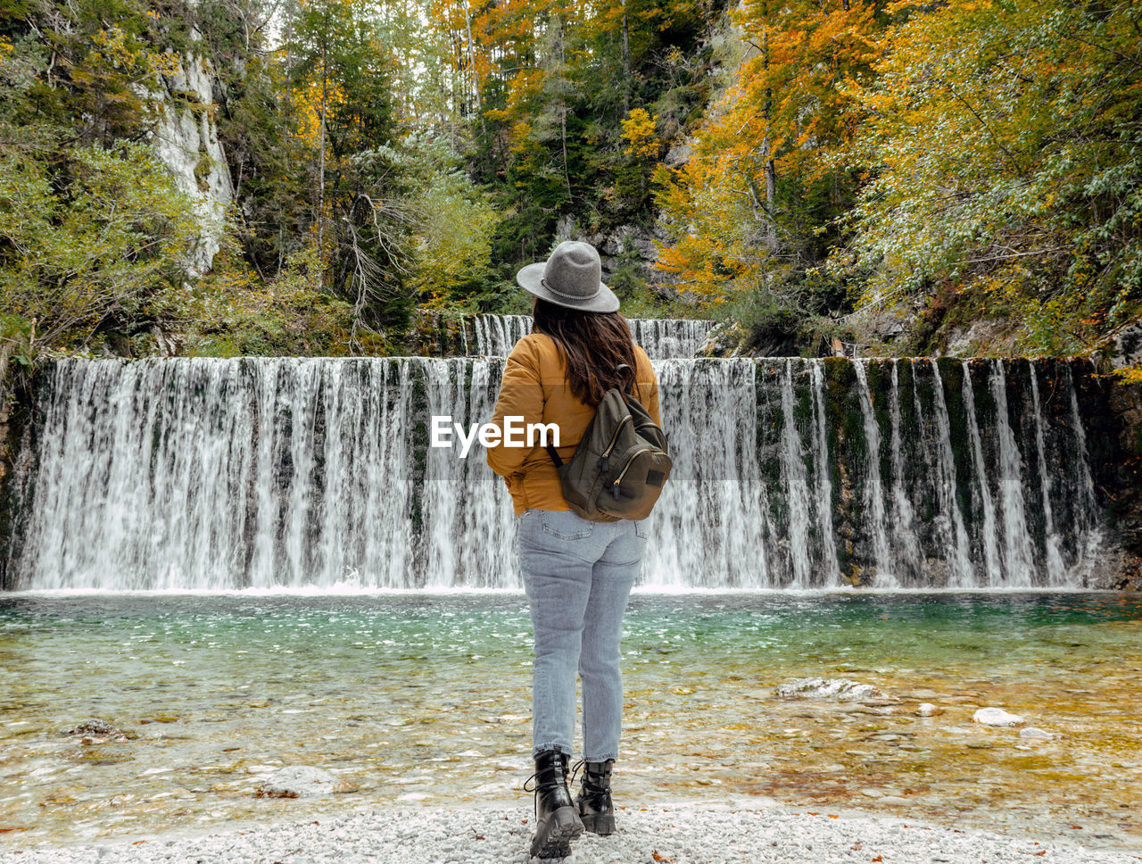 waterfall, water, one person, full length, beauty in nature, motion, tree, nature, water feature, leisure activity, flowing water, autumn, scenics - nature, plant, standing, lifestyles, clothing, adult, day, casual clothing, young adult, rear view, hat, long exposure, women, forest, body of water, outdoors, spring, footwear, flowing, travel, vacation, tourism, fashion accessory, trip, land, splashing, non-urban scene, walking, person, river, holiday, activity, environment, travel destinations, hiking, blurred motion, backpack, idyllic