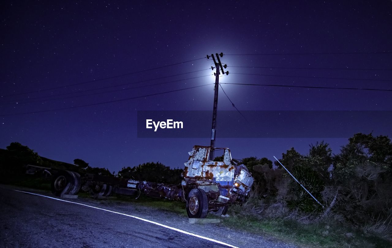 CARS ON ROAD BY ILLUMINATED FIELD AGAINST SKY AT NIGHT