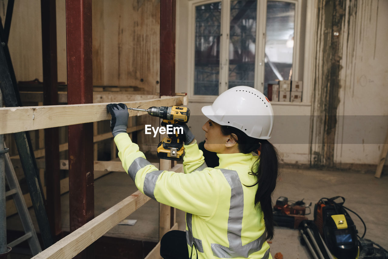Female worker in hardhat using drill on wooden plank while working at construction site