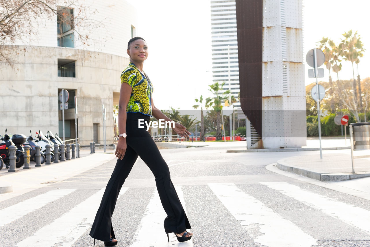 Side view full body of self assured young fashionable african lady with short hair in trendy outfit and high heels smiling and crossing road in city
