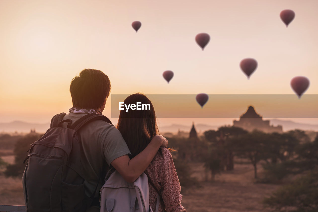 Rear view of couple standing against sky looking at hot air balloon during sunset