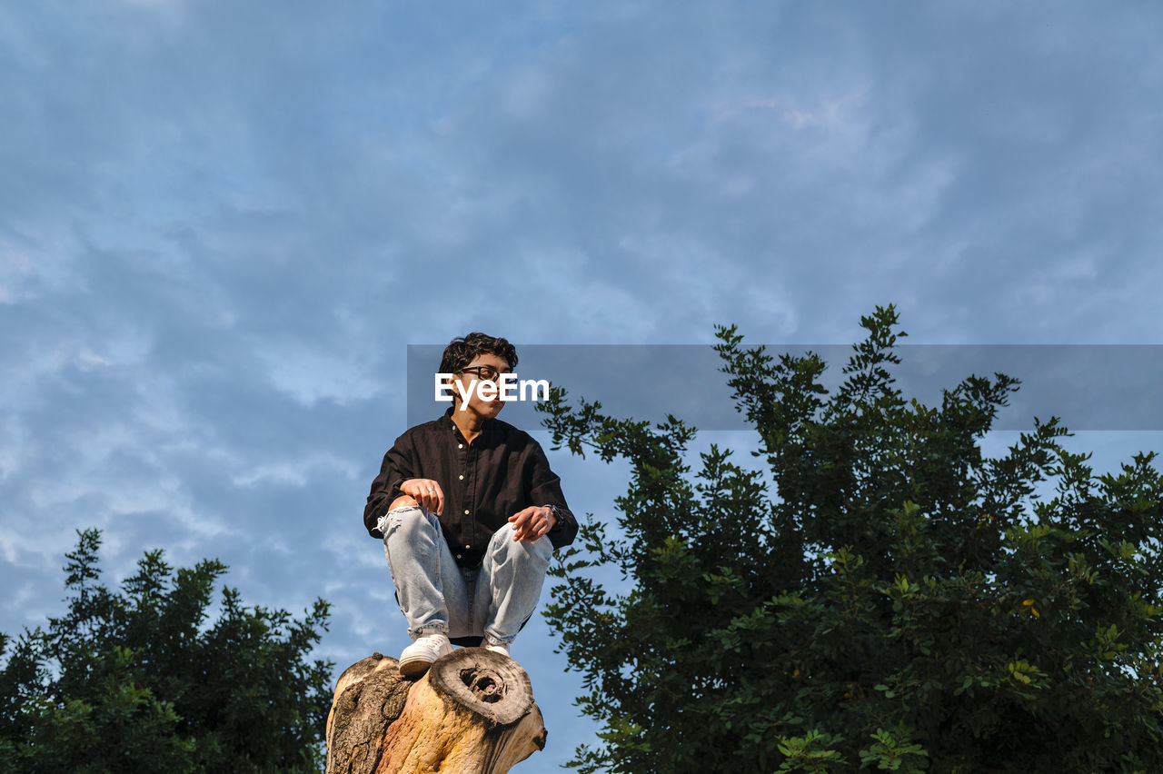 Young transgender man with glasses posing on a log outdoors.