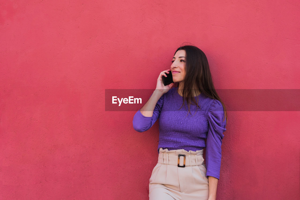 Positive young female in violet blouse and light beige pants talking on mobile phone while standing against colorful red wall background