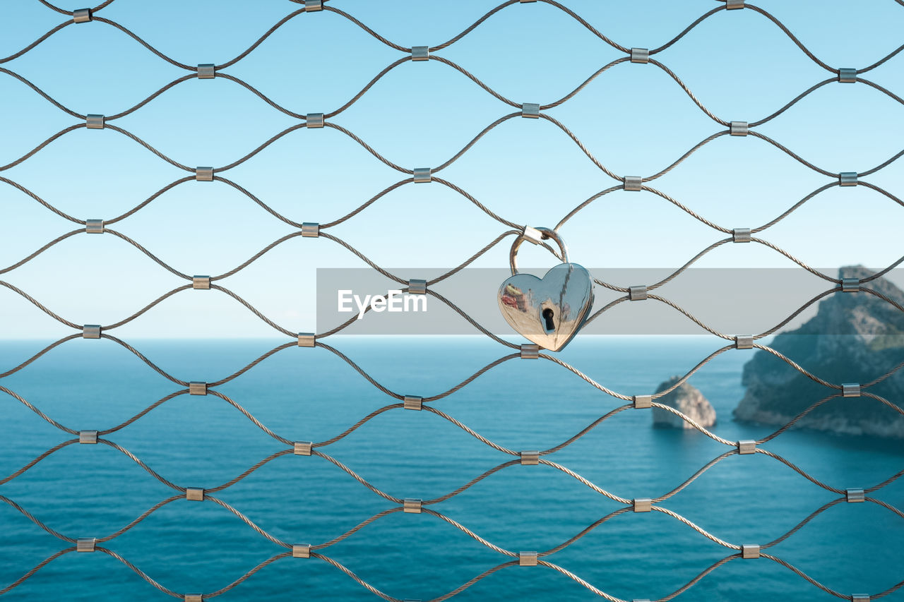 Heart shape love lock hanging on fence against mediterranean sea