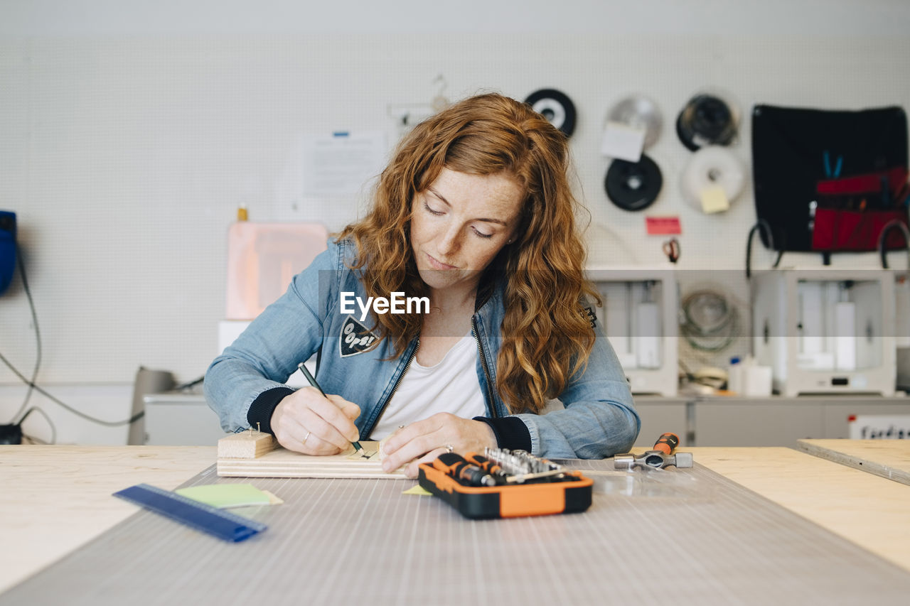 Confident redhead female engineer writing on wood at workbench in creative office