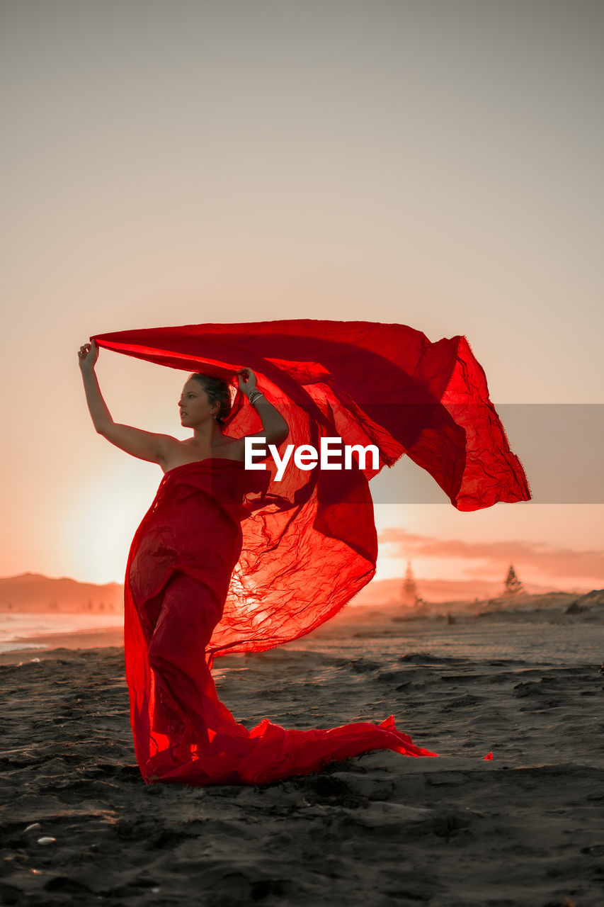 Full length of woman wearing red dress standing in beach against sky during sunset