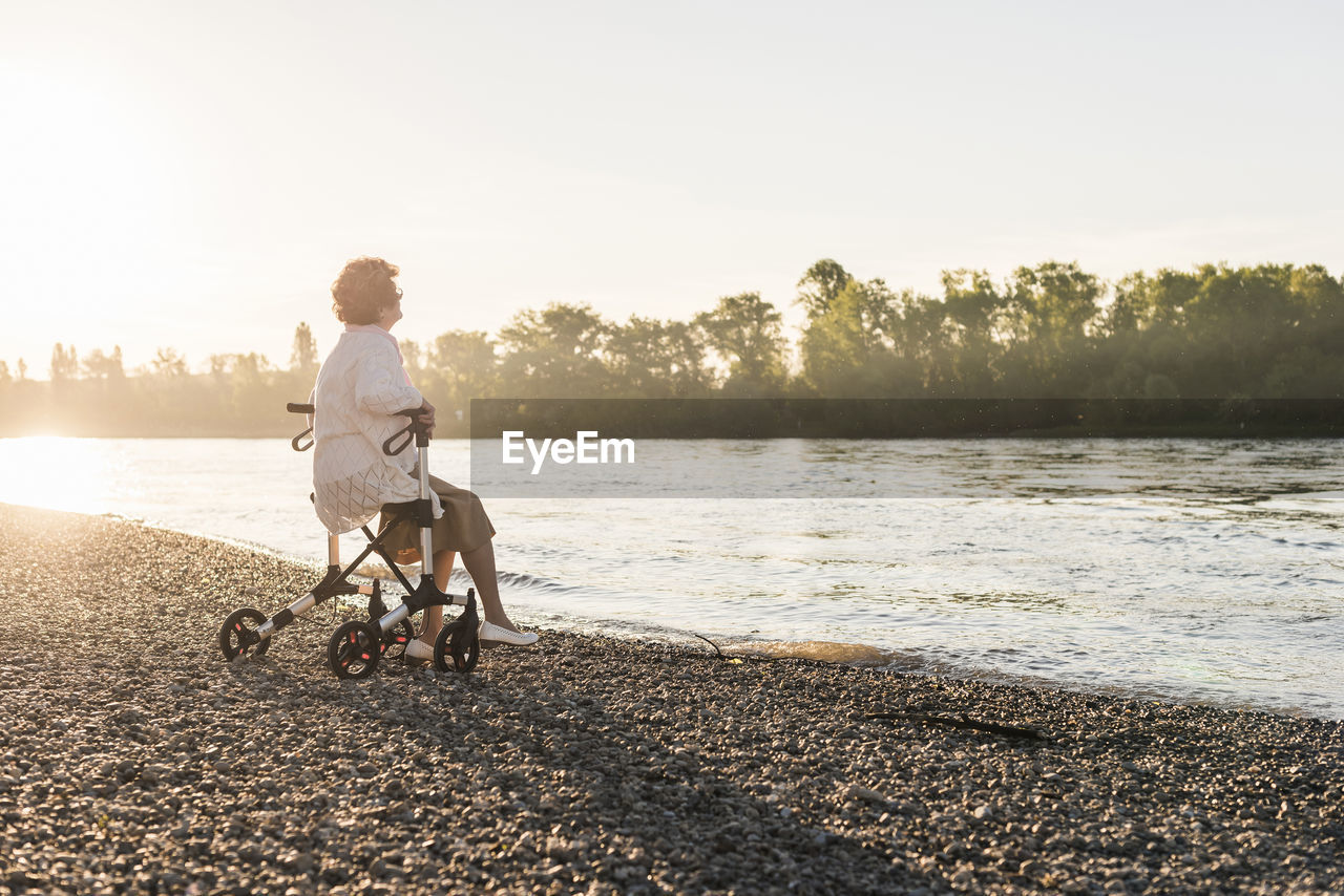 Pensive senior woman sitting on her wheeled walker at riverside