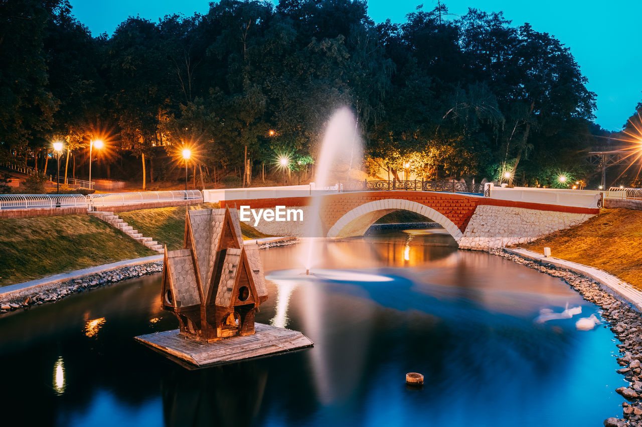 ILLUMINATED FOUNTAIN BY LAKE AT NIGHT