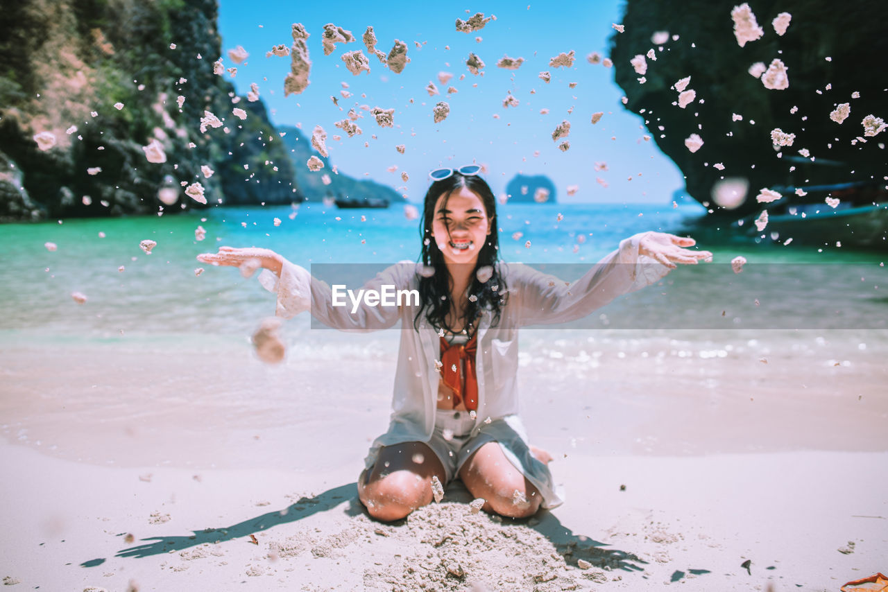 Smiling young woman throwing sand at beach during sunny day