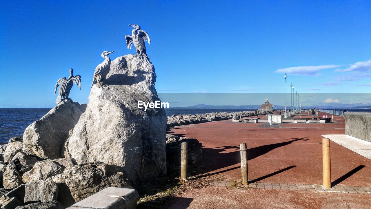 BIRDS PERCHING ON ROCK BY SEA AGAINST SKY