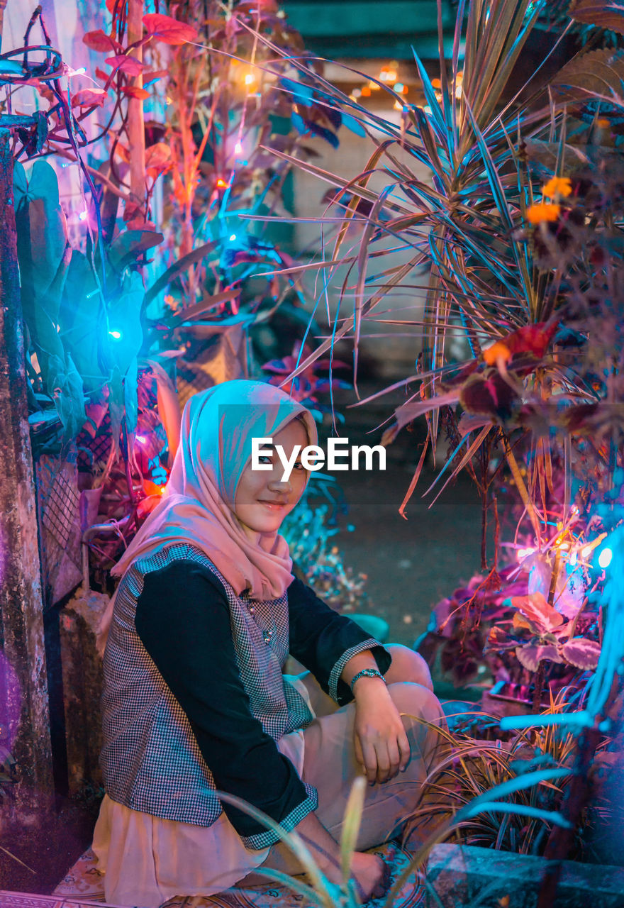 Portrait of smiling young woman wearing hijab sitting by illuminated decorations at night