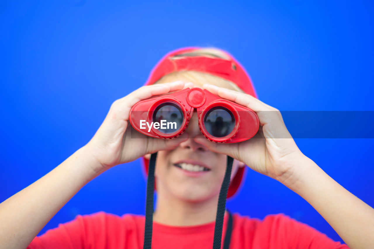 Close-up of boy looking through binoculars while standing against blue background