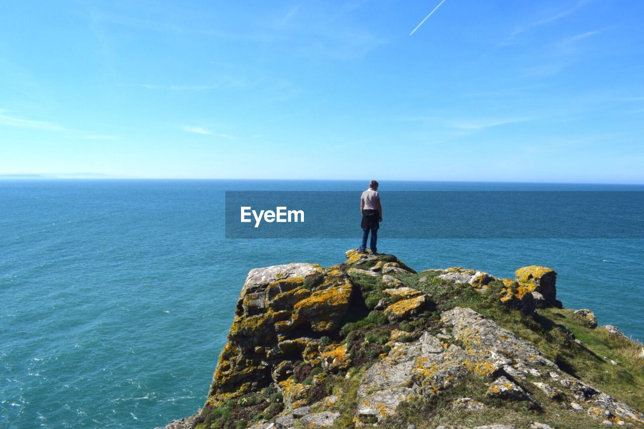 Rear view of mature woman looking at sea while standing on mountain against blue sky