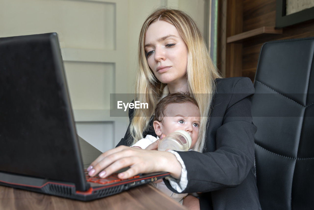 Young businesswoman holds baby in her arms, in office, combining this with working on laptop