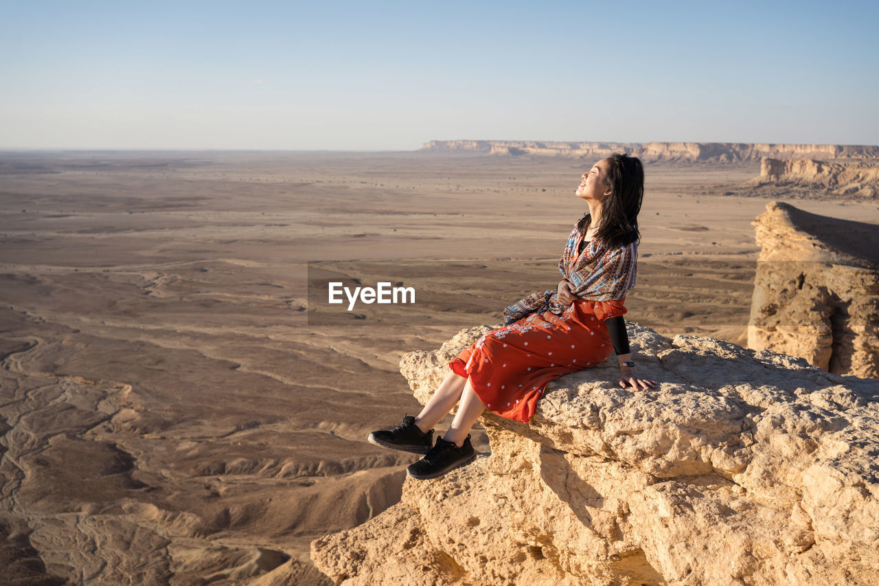 Side view of ethnic asian woman in casual clothes sitting on cliff with closed eyes contemplating the views in the edge of the world in saudi arabia