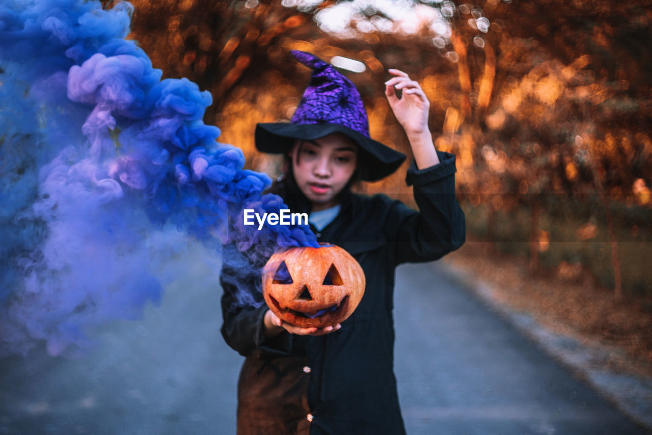 Young woman holding jack o lantern emitting smoke while standing on road during halloween