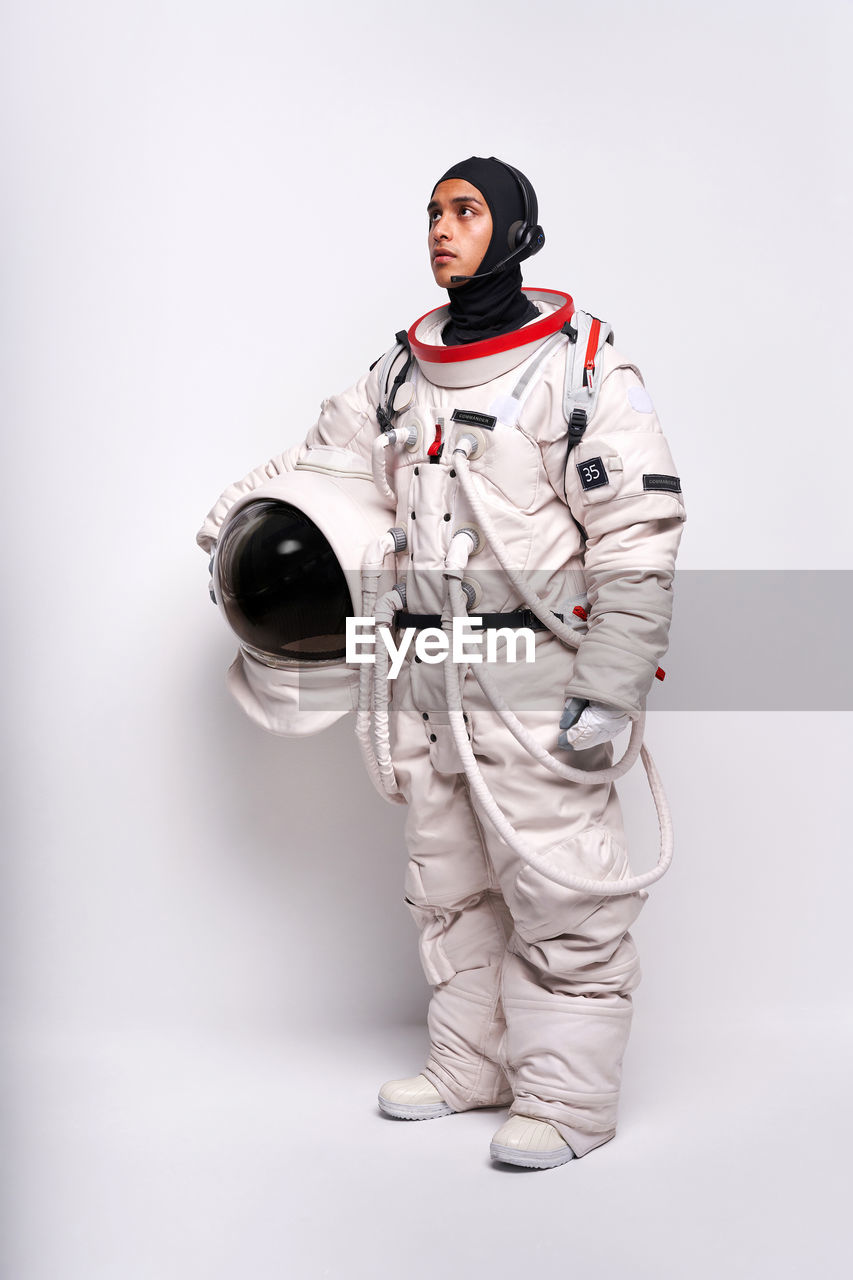 Full body of colombian male astronaut in spacesuit with helmet in hand standing in studio against white background and looking away