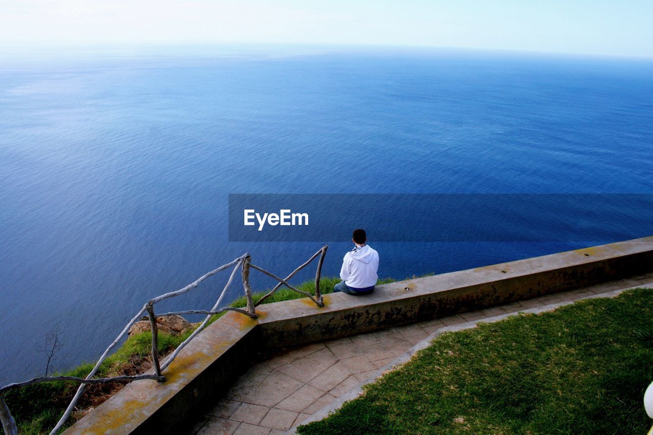 Rear view of man sitting on retaining wall against blue sea