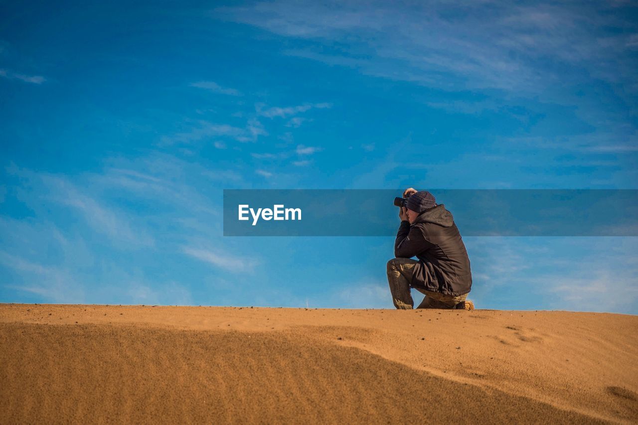 Side view of man photographing with digital camera in desert against sky