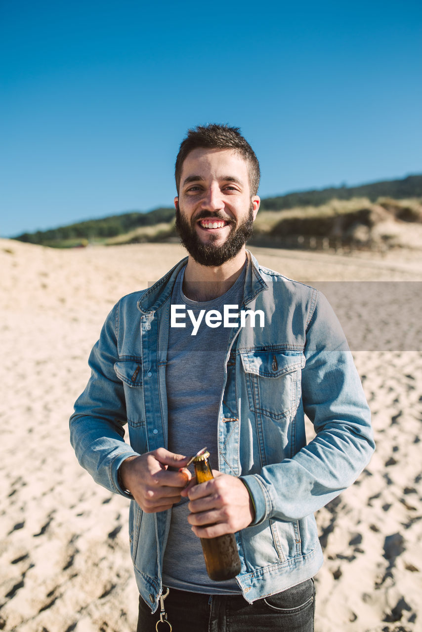 Portrait of young man opening bottle while standing on sand