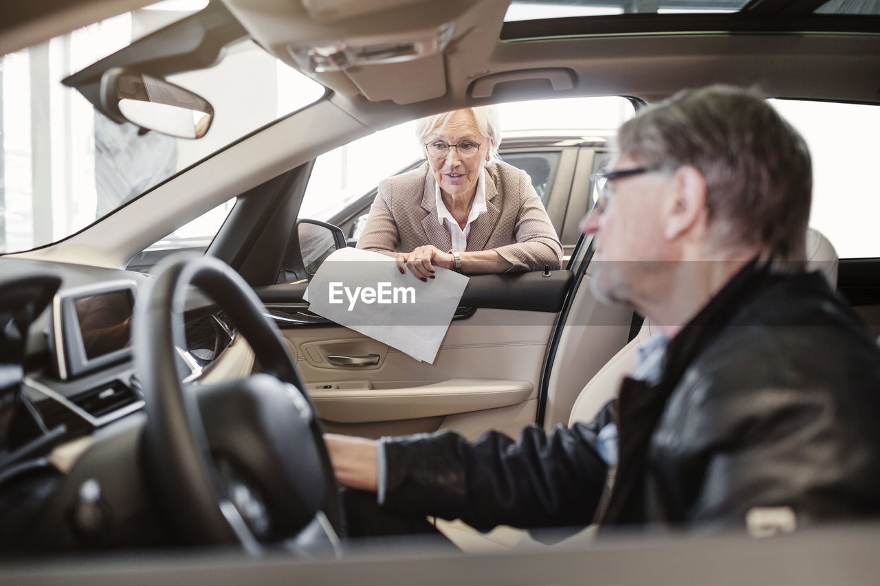 Senior saleswoman explaining features to man sitting in car at shop