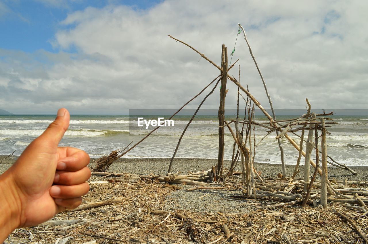 Human hand forming ok sign before broken wooden structure on beach against cloudy sky