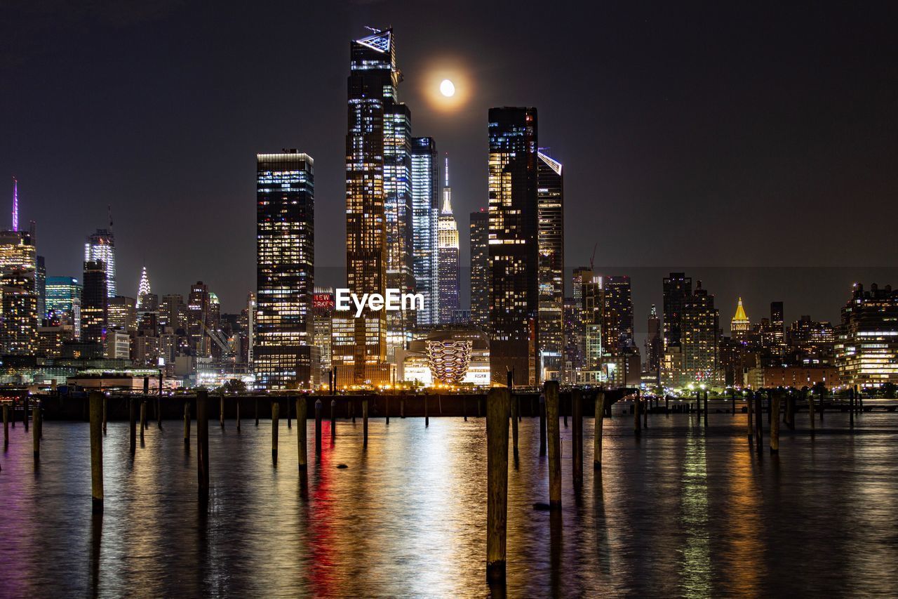 Illuminated buildings by river against sky at night