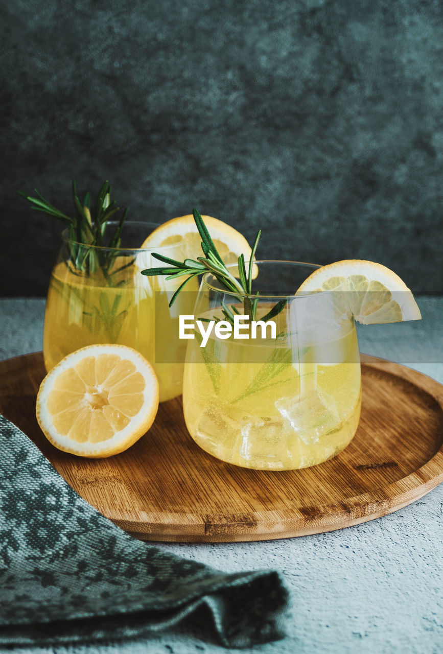 Glasses with cold cocktail, lemonade, sliced lemon and rosemary plant on the concrete table