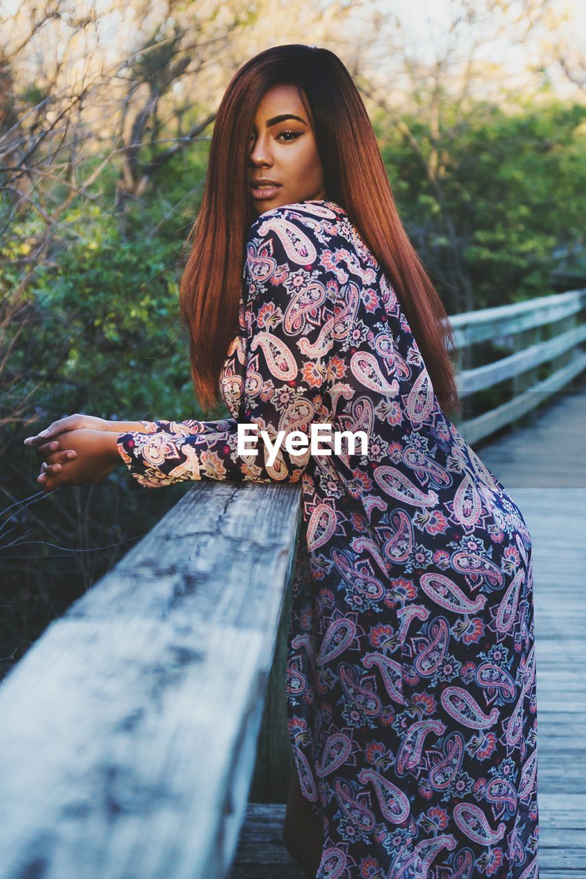 Portrait of young woman leaning on railing at wooden footbridge by plants