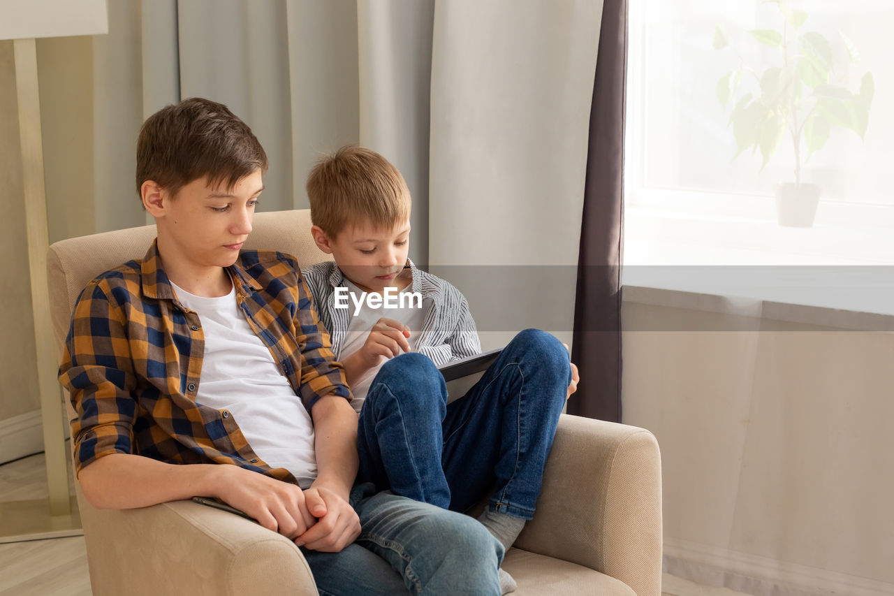 Two boys,  sit in beige armchairs, in a room, during the day, using mobile device