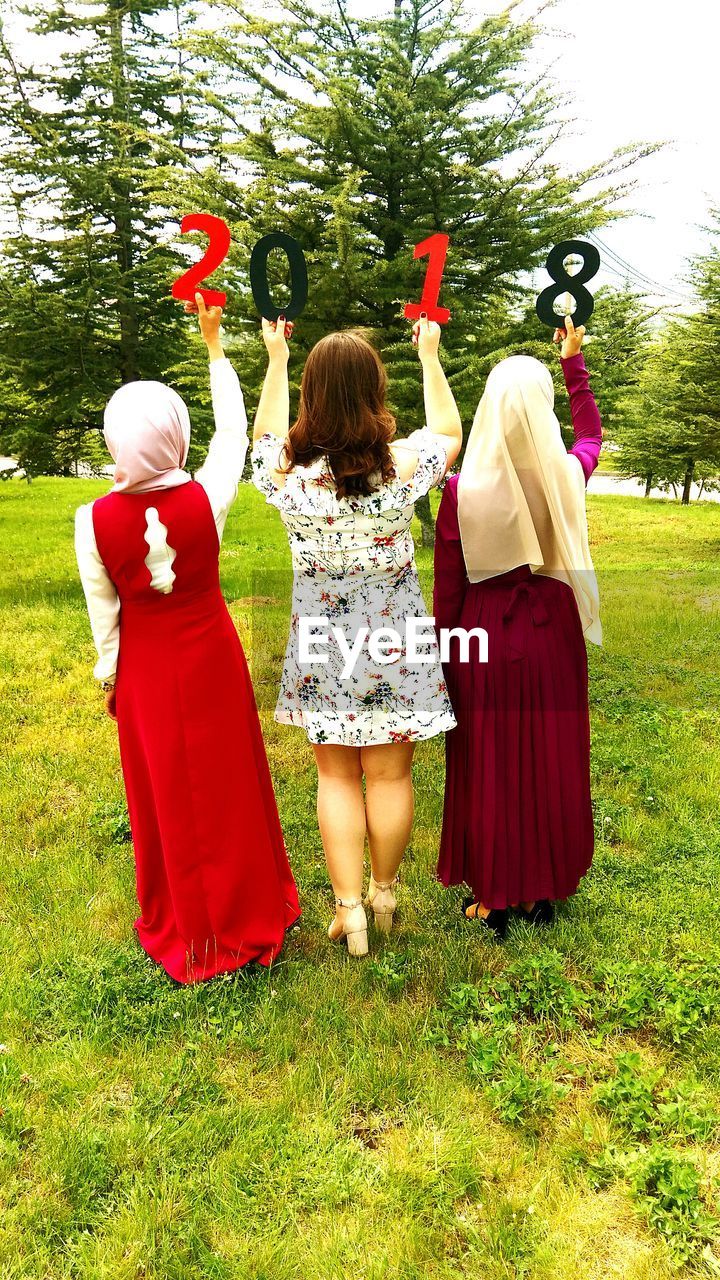 Rear view of women with arms raised holding numbers while standing on field at park